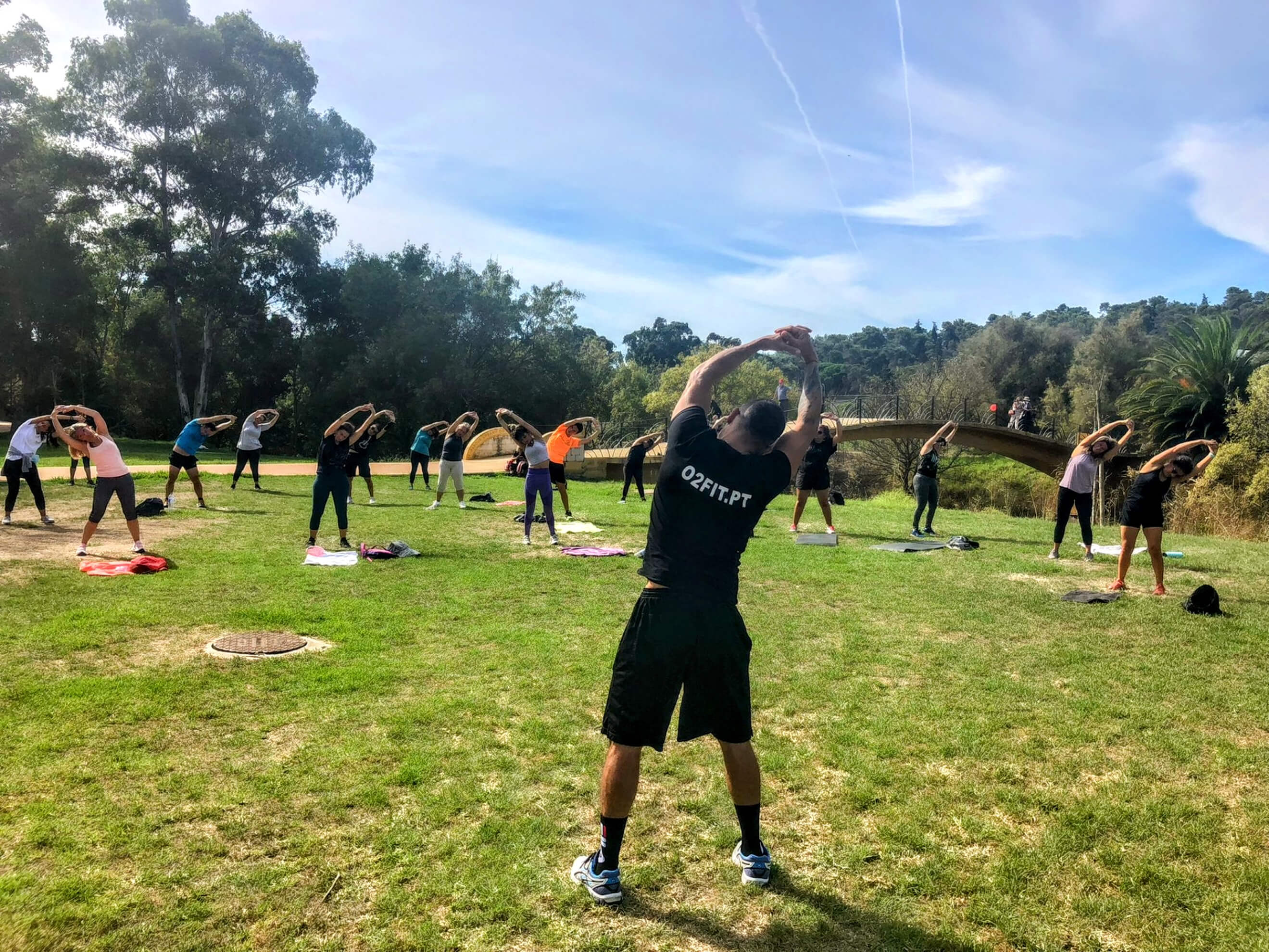 Grupo O2Fit a treinar ao ar livre num relvado, de braços no ar